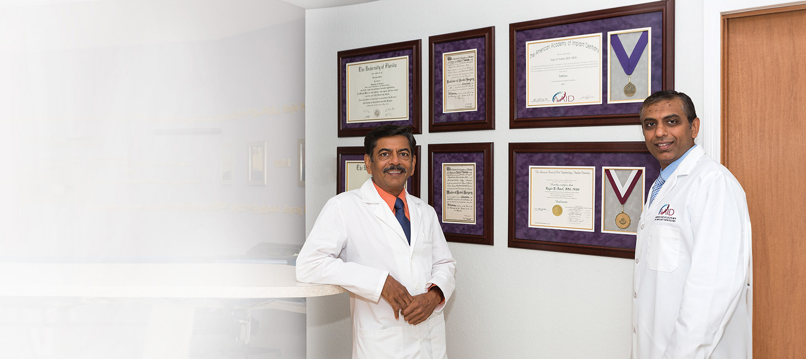 The image shows two individuals, one standing and the other sitting, both wearing white lab coats, in front of a wall displaying multiple framed certificates. They appear to be in an office setting with professional decor.
