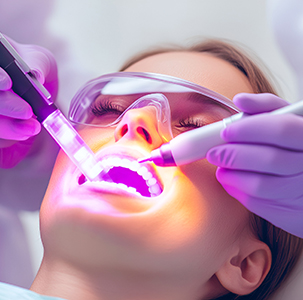 The image shows a woman lying down in what appears to be a dental or medical setting, receiving treatment on her teeth using a device that emits purple light.