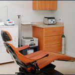 The image shows an interior view of a dental office with a modern dental chair, a computer monitor on a countertop, and various medical equipment.