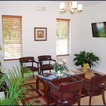 The image shows an interior view of a waiting area with a television screen mounted on the wall, displaying static content, and a reception desk with chairs in front of it. There are two potted plants, one near each window, and a decorative vase on the counter.
