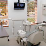 The image shows a dental office interior with a modern chair, a desk with a computer monitor, and various dental equipment.