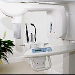 The image shows an interior view of a medical facility, featuring a large, modern dental chair with a digital panel and a ceiling-mounted overhead light.