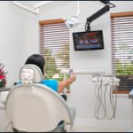 The image depicts an interior dental office setting with a woman seated in a dental chair, facing the camera, holding a smartphone.
