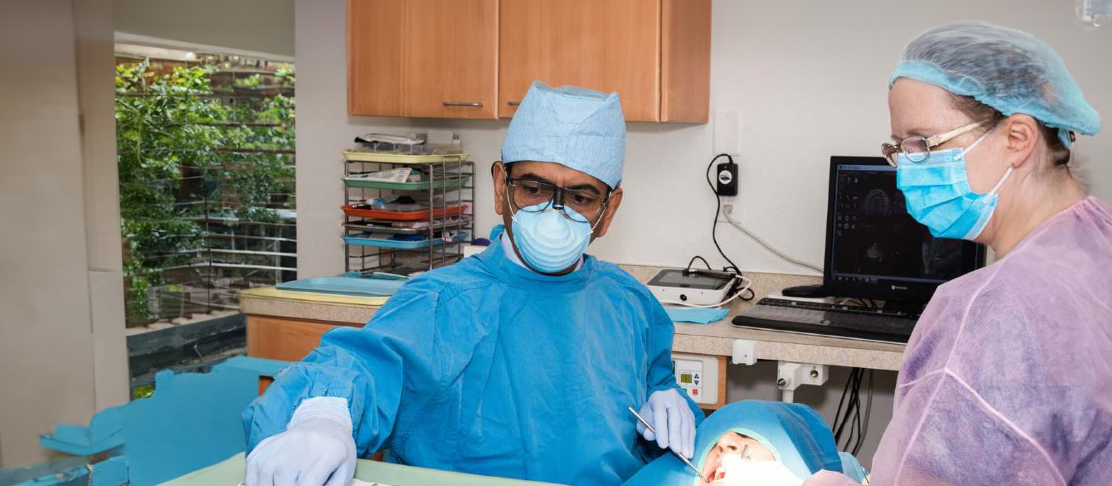 The image shows a person in a medical gown and surgical mask standing next to a table with medical equipment, while another individual, likely a healthcare professional, is sitting at the table.