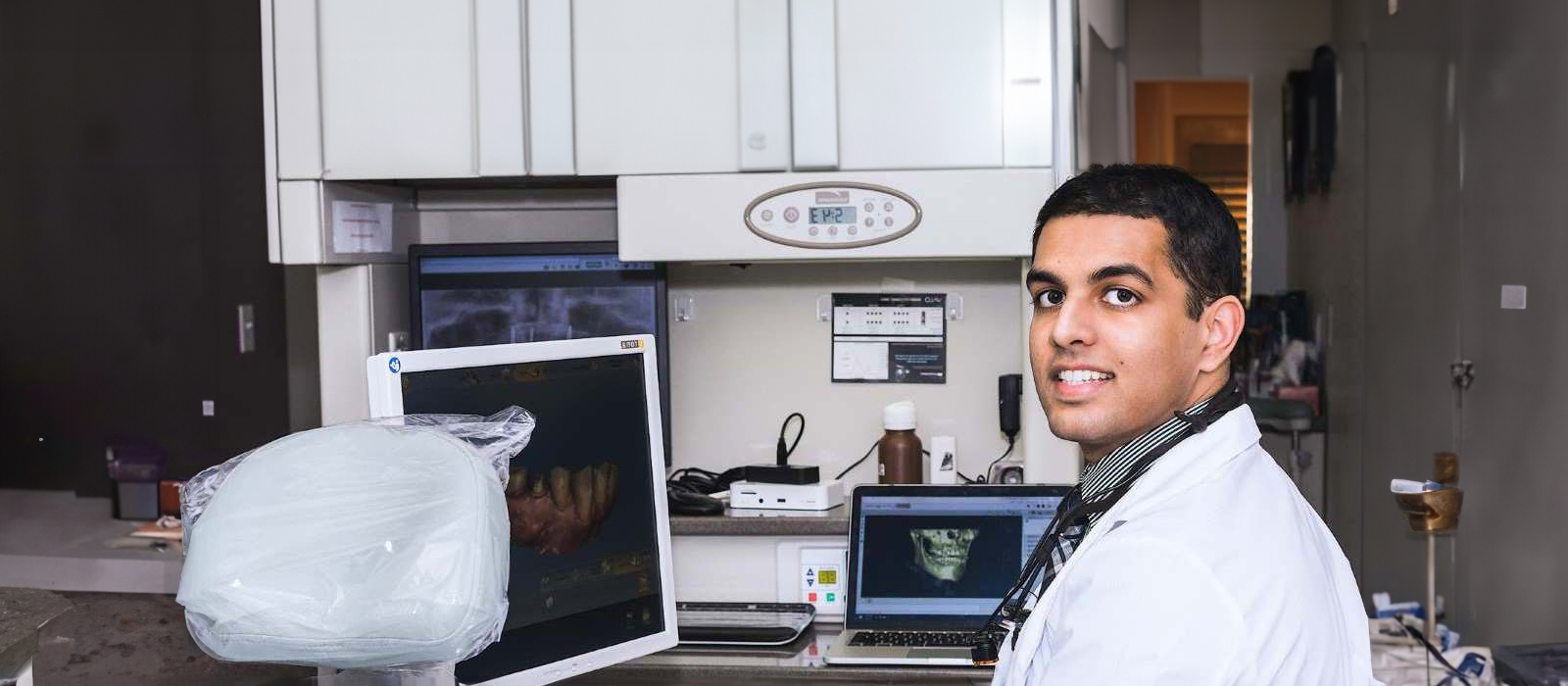 The image shows a man in a lab coat standing next to a piece of medical equipment, likely an MRI machine, with a smile on his face.