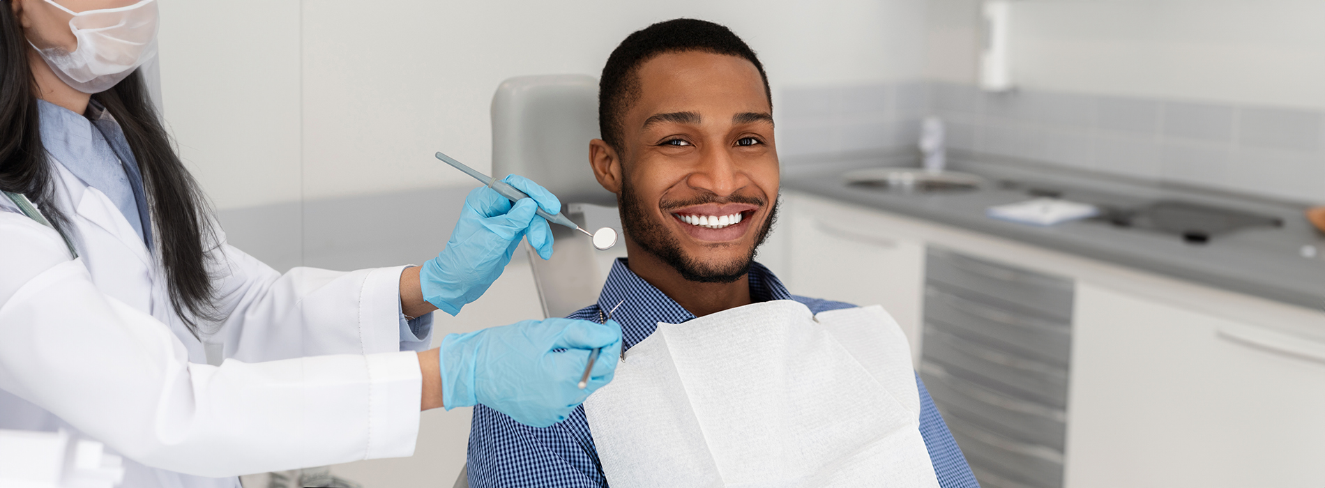 A dental hygienist is assisting a smiling man in a dentist s chair, with dental tools and equipment visible.