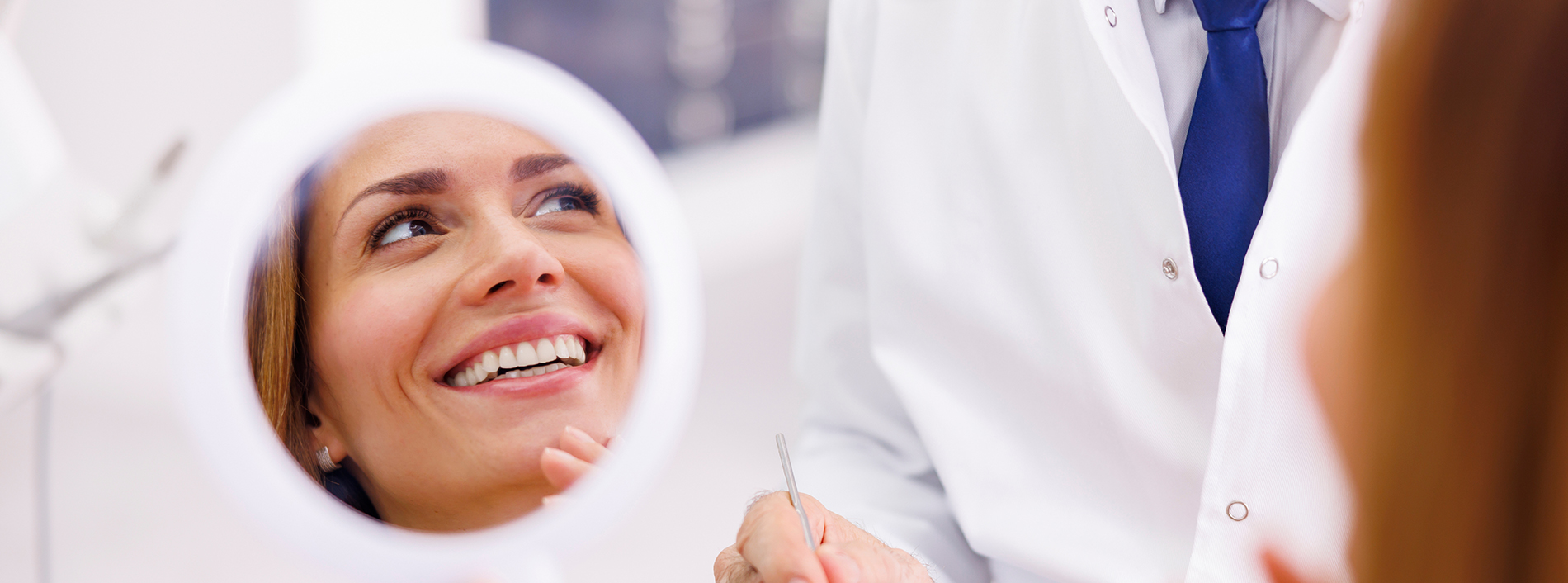 A smiling woman sitting in front of a mirror, being attended to by a dental professional.