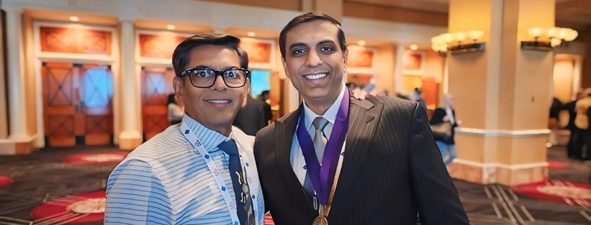 The image shows two men standing side by side, one wearing a suit and tie with a medal around his neck and the other in a casual shirt. They are indoors, likely at an event, as suggested by the background that includes a stage and decorative elements.