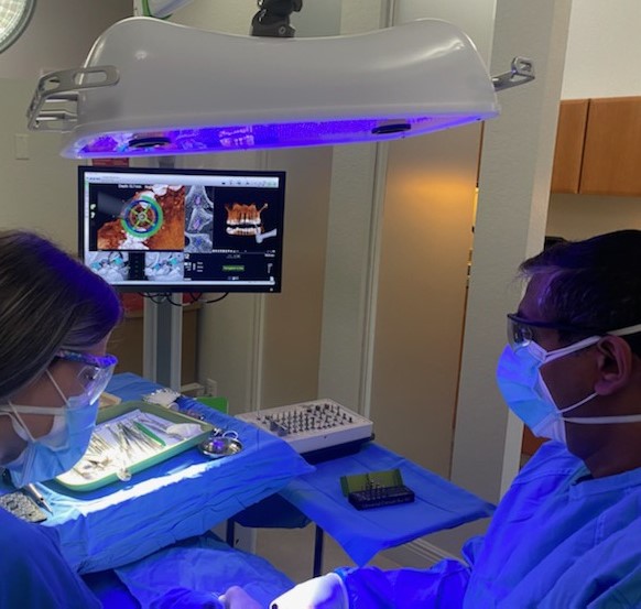 A person in a surgical mask and gown is operating an ophthalmic machine, with a monitor displaying a medical image, assisted by another individual in a similar setting.