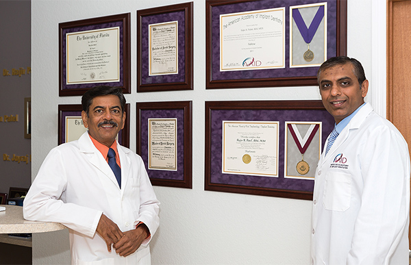 The image features two individuals, one wearing a white lab coat and the other in traditional Indian attire, standing side by side in front of framed certificates on a wall. They are likely professionals in an academic or medical setting.