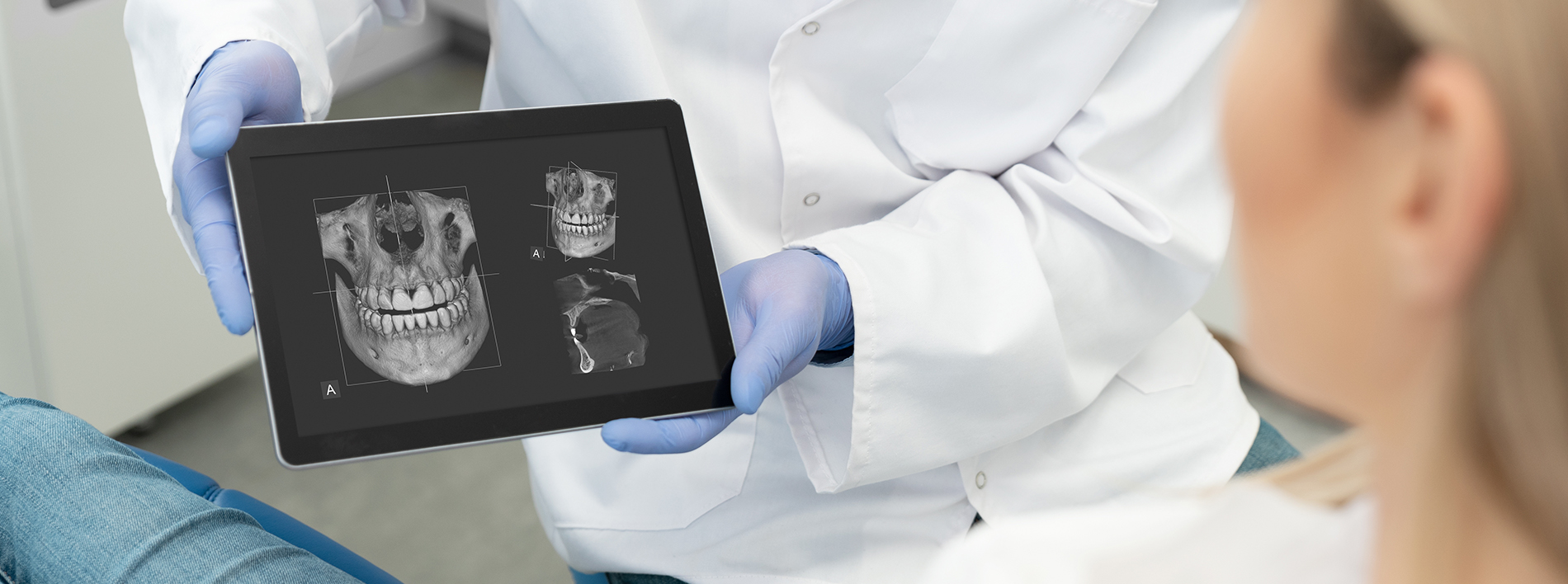 A dental professional in a lab coat holding up an iPad displaying a 3D model of a human skull to a seated patient.