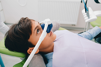 A young person sitting in a dental chair, wearing an eye shield, with their head tilted towards the camera.