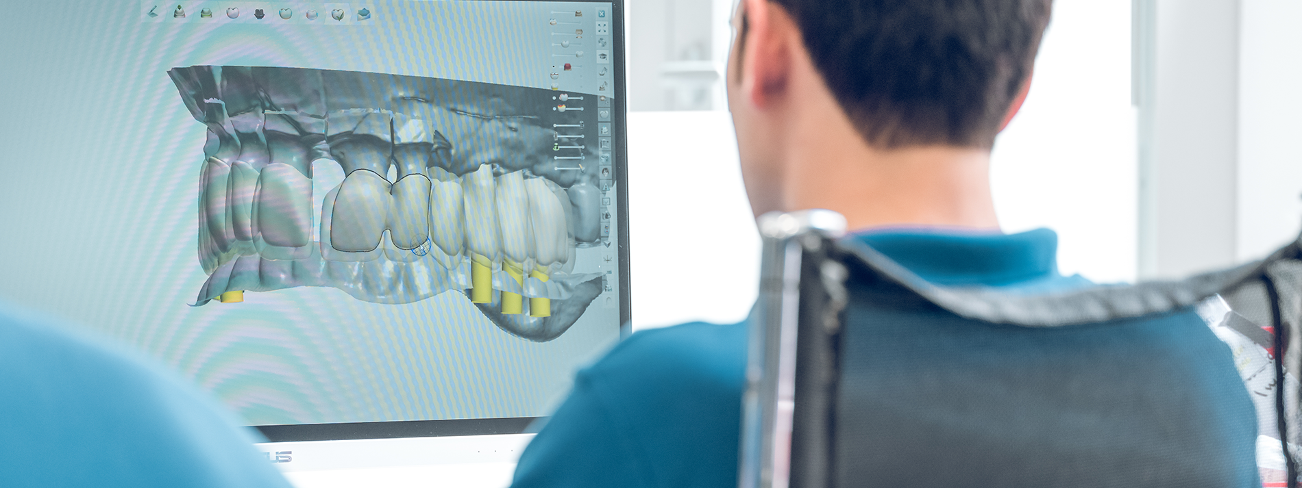 The image is a photograph showing two individuals engaged in an activity involving a large screen displaying 3D imagery of a dental implant and surgical tools, with one person seated at the screen while another stands behind them, both focused on the content. They appear to be in a professional setting, possibly a dental or medical facility, with a clean and sterile environment.
