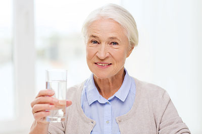 The image shows an elderly woman holding a glass of water and smiling, with the background blurred but suggesting an indoor setting.