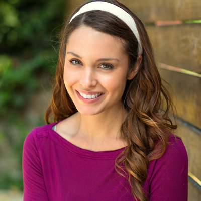 A woman with a smile, wearing a purple top and headband, poses for the camera against a wooden fence.