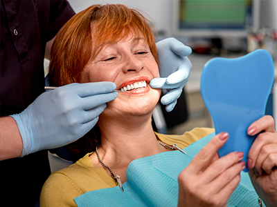 A woman in a dental chair receiving a teeth cleaning, with a hygienist holding a mirror and blue paste on her teeth.