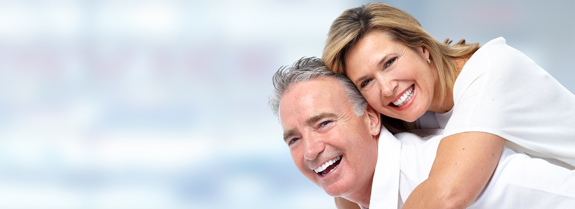 An elderly couple smiling and hugging, with the man wearing a white shirt and the woman in a light-colored top, set against a blurred background.