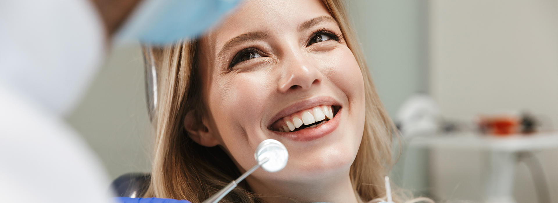 The image shows a young woman in a dental office, smiling at the camera while seated in an examination chair. She appears to be undergoing a dental procedure or consultation, with a dental professional visible behind her.