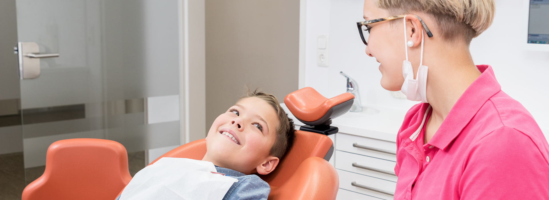 A dental office scene with a dentist and patient in the foreground, an assistant in the background, and a smiling child seated on a chair.