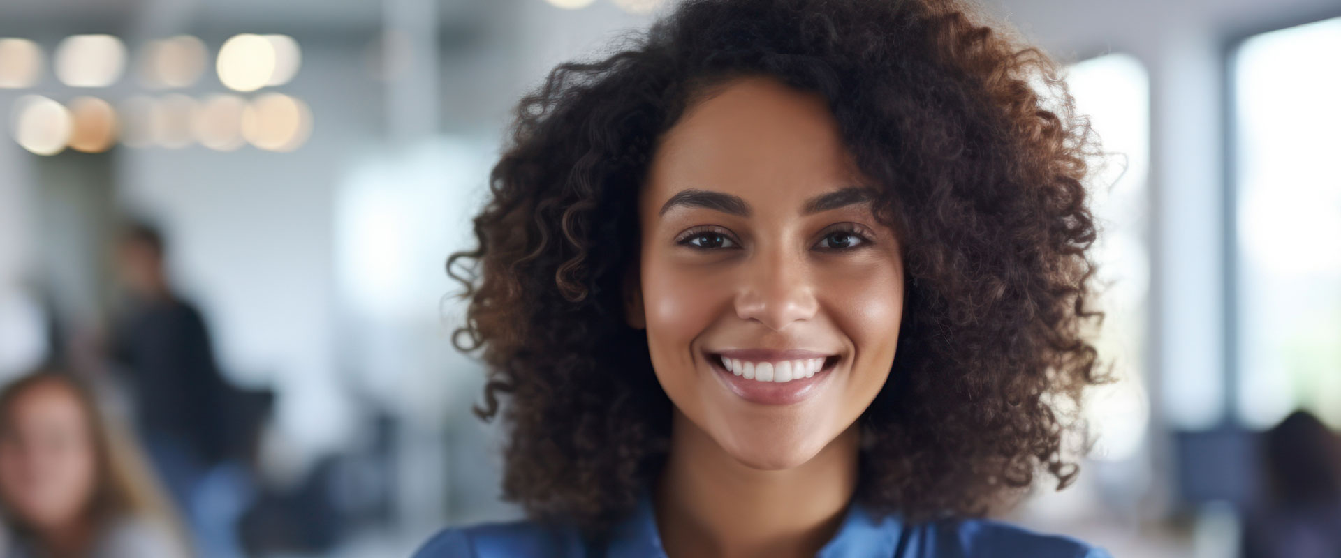 A woman with a warm smile, wearing business attire, stands confidently in an office environment.