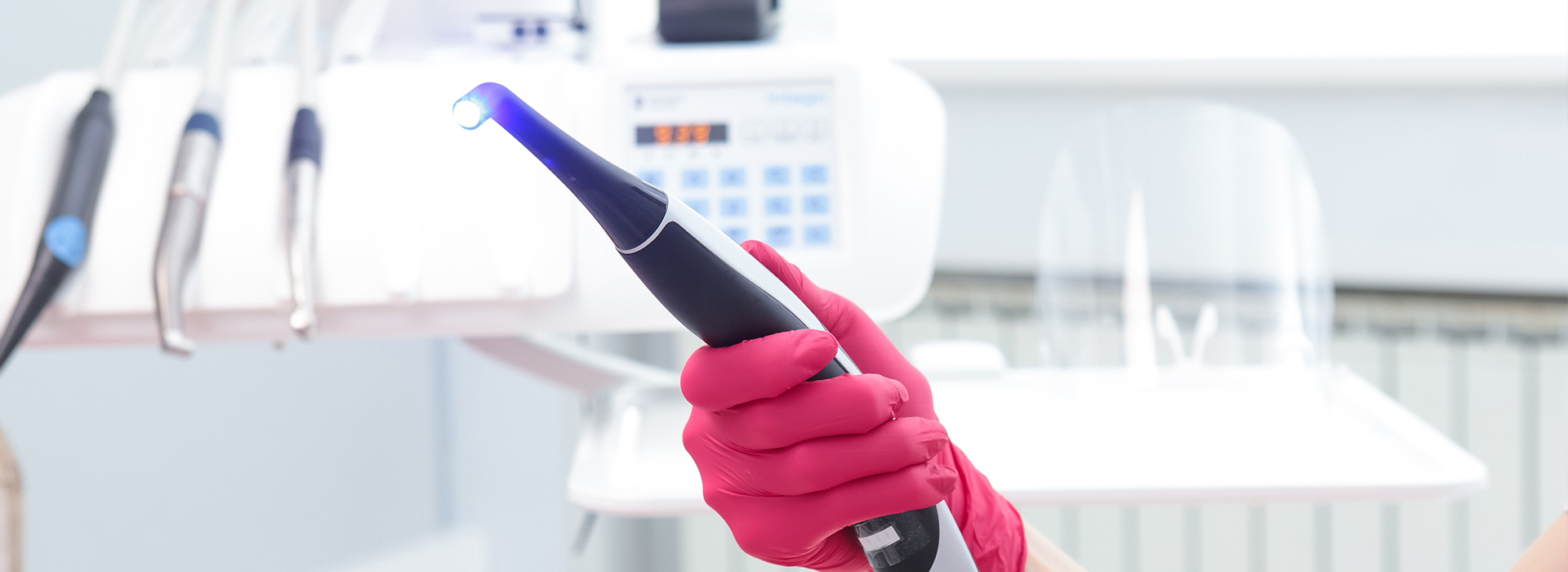 The image shows a person wearing pink gloves and holding a blue device, which appears to be a handheld ultrasound machine, in front of their face. They are standing in a room with white walls, and there is medical equipment in the background, suggesting a clinical or healthcare setting.