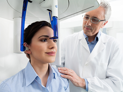 The image depicts a medical setting with a person seated in front of a 3D imaging device, receiving a scan or examination. A medical professional stands beside the individual, observing the procedure.