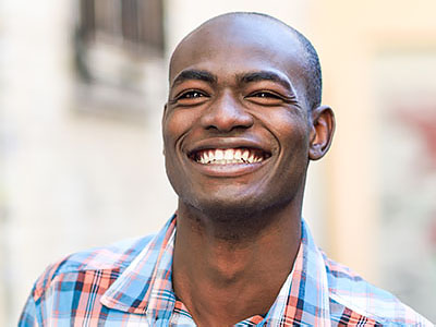 A smiling man with short hair, wearing a plaid shirt, stands outdoors in front of a building.