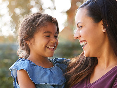 The image shows a woman and a young child smiling at the camera, with the woman holding the child. They appear to be outdoors during daylight.