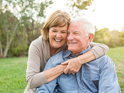 A man and a woman are affectionately embracing each other outdoors, with the man wearing glasses and both individuals appearing to be elderly.