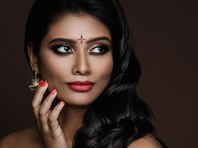 A photograph of a woman with dark hair, wearing red lipstick and a red nail on her index finger. She is dressed in a black top with a scoop neckline and has a neutral expression while looking slightly to the side.