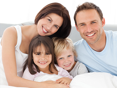 The image is a photograph of a family, likely taken in a domestic setting. It features a man and woman with three children sitting on a bed.