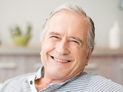 The image shows an elderly man with gray hair, wearing a blue shirt and a white beard. He is smiling and appears to be sitting in a comfortable environment, possibly his home.