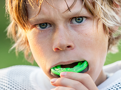 The image shows a young boy with blonde hair, wearing a football uniform and holding a green mouthguard in his mouth. He is looking directly at the camera with a serious expression.