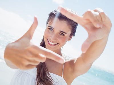 A woman with long hair, smiling and holding up a finger in front of her face against a beach backdrop.