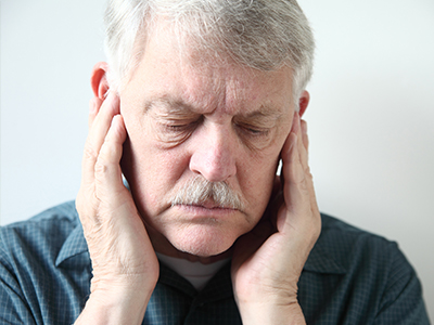 The image shows an older man with his eyes closed, holding his face with both hands, and appears to be in a state of distress or discomfort.